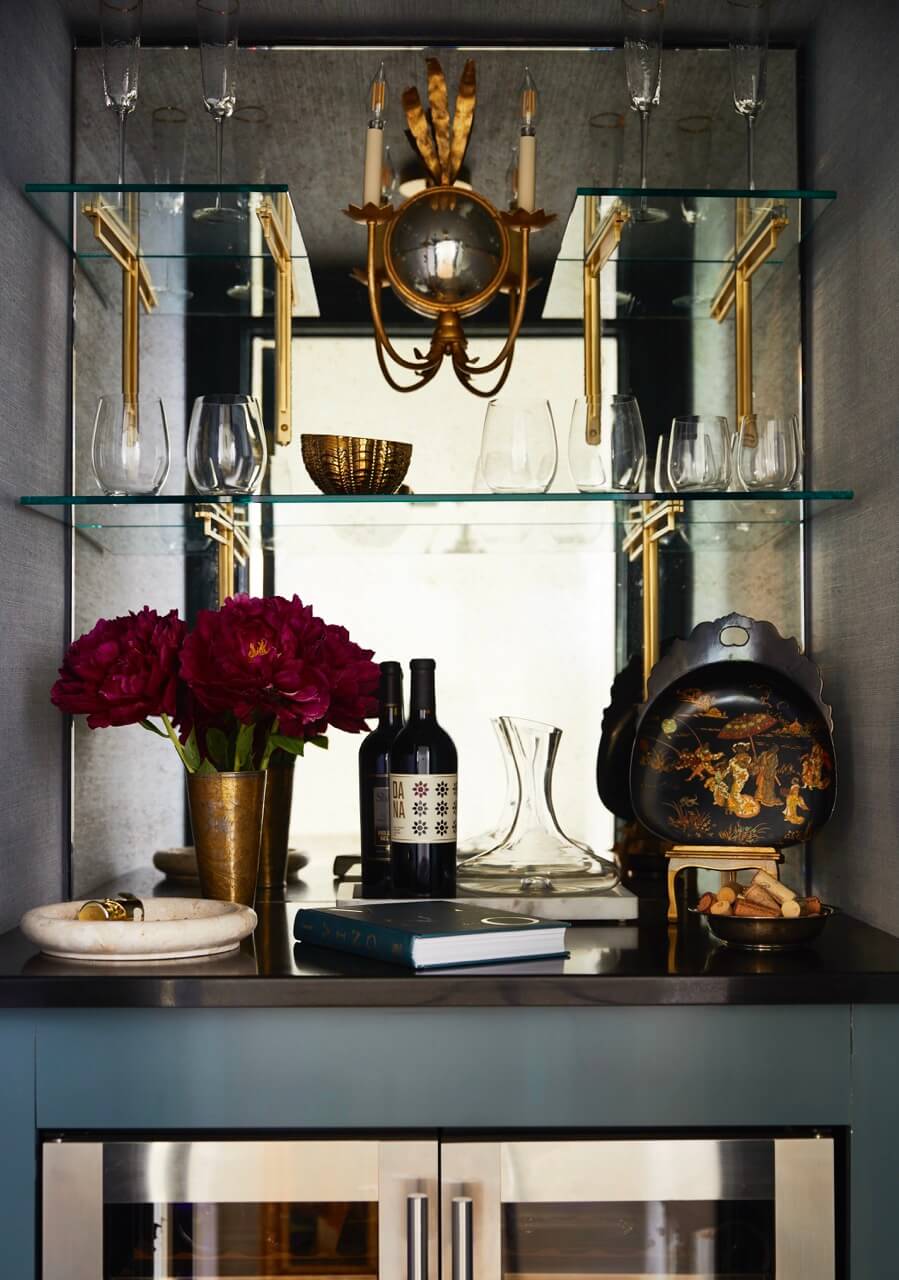 A home bar with wine glasses, decanter, bottles, red flowers in a gold vase, decorative tray, book, and mirrored shelves under a wall sconce with gold accents.