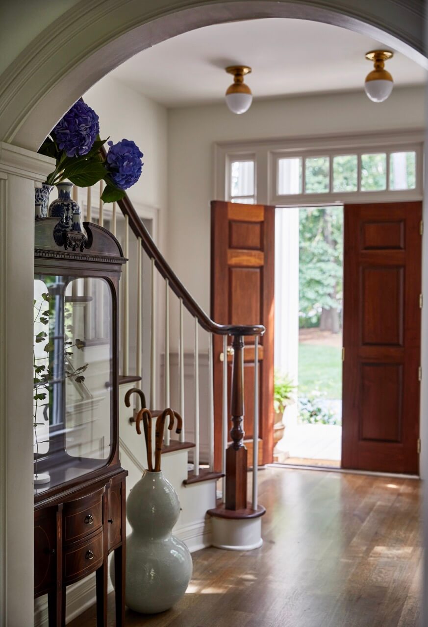 A bright hallway with wooden floors, an open front door, a staircase, and a glass display cabinet evokes lakeside beauty, featuring vases holding purple flowers and umbrellas in this inviting South Carolina retreat.