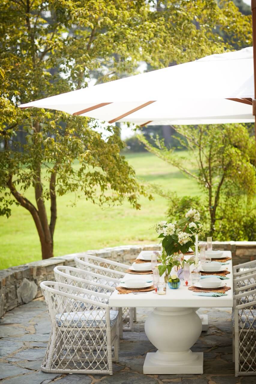 A white outdoor dining table with wicker chairs sits under a large umbrella, surrounded by greenery and trees on a stone patio—perfect for enjoying lakeside beauty at this South Carolina retreat.