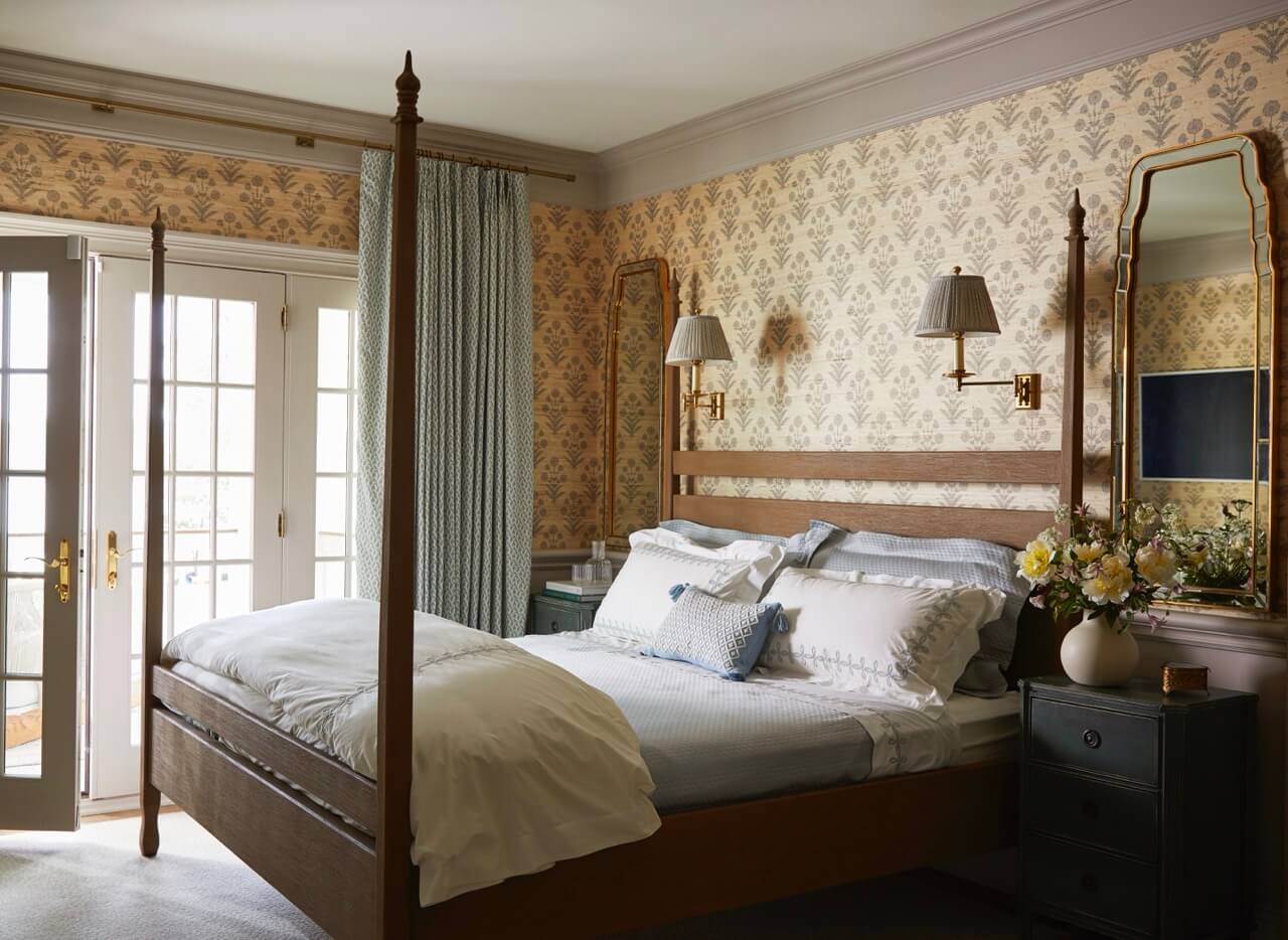 A neatly made bed with white linens and patterned pillows sits between two nightstands in a classic bedroom with floral wallpaper, mirrors, and French doors letting in natural light at this charming South Carolina retreat.