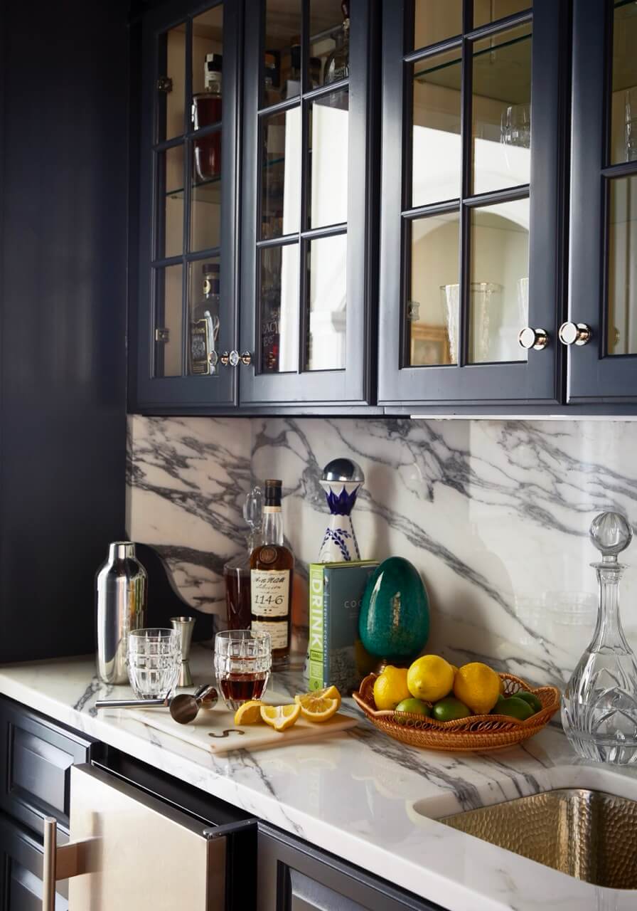 A butler's pantry with a marble backsplash displays liquor bottles, glassware, a cocktail shaker, and a basket of lemons and limes beneath dark glass-front cabinets in this charming South Carolina retreat.