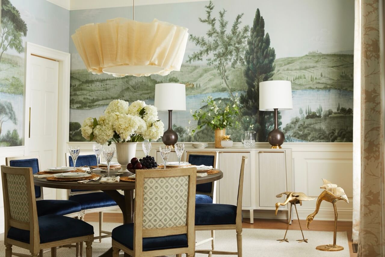 Elegant dining room with round table, blue upholstered chairs, and floral centerpiece captures lakeside beauty, while a large landscape mural and sideboard with lamps evoke the charm of a South Carolina retreat.