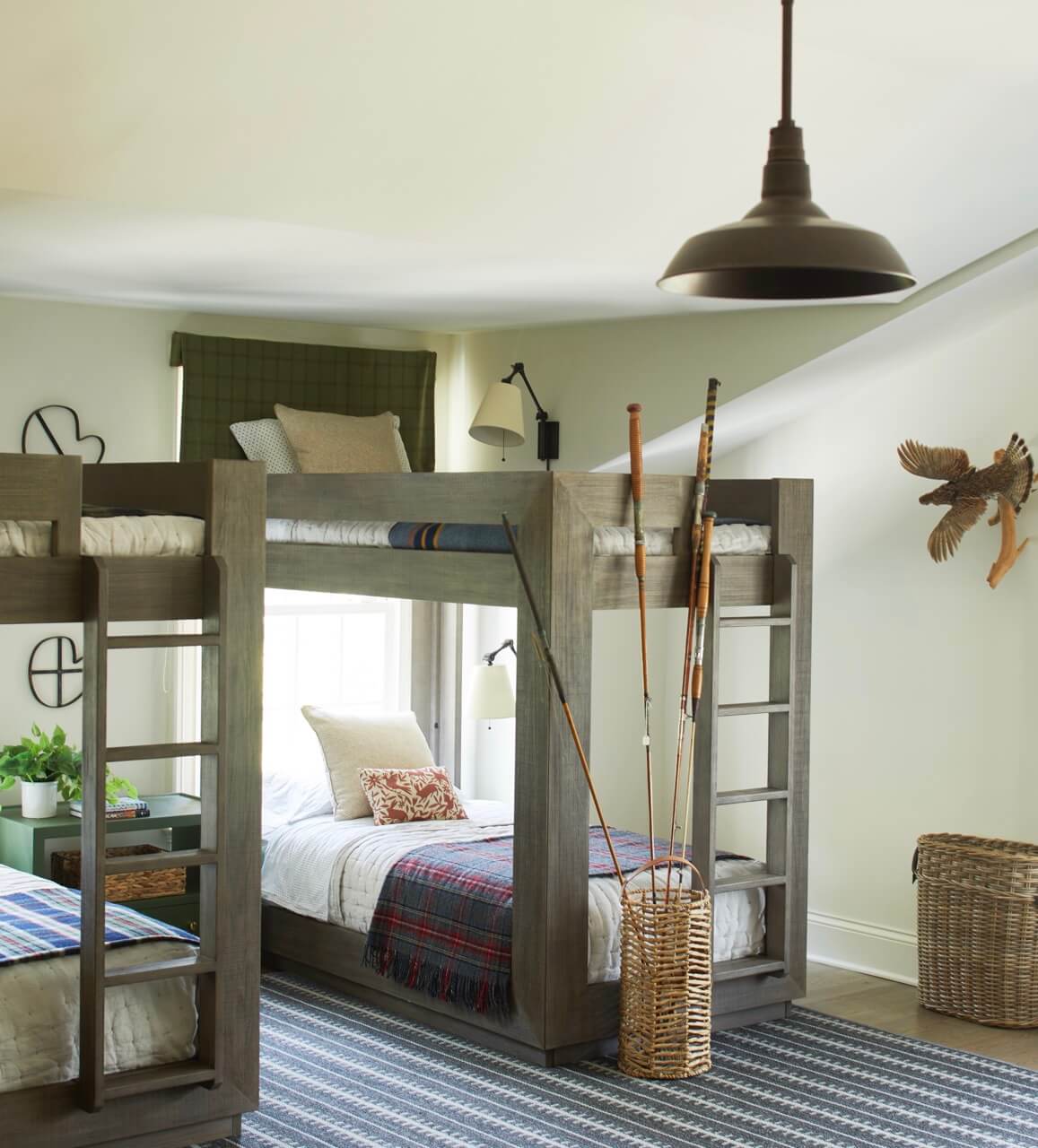 A cozy South Carolina retreat, this bedroom features two sets of wooden bunk beds, plaid bedding, a striped rug, a wicker basket holding hockey sticks, and a window with a green valance overlooking lakeside beauty.