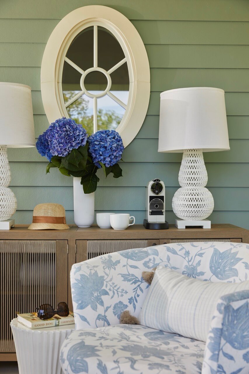 A wooden sideboard with two white lamps, a coffee machine, blue hydrangeas in a vase, and a round mirror against a green wall; evoking lakeside beauty at a South Carolina retreat. A floral armchair and hat are in the foreground.