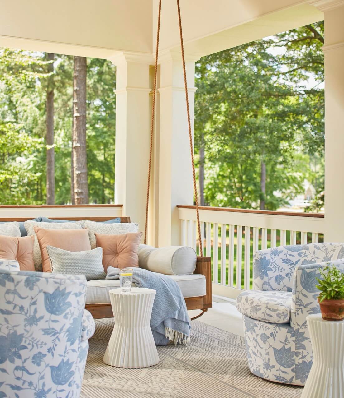 A covered porch with a wooden swing sofa, blue and white floral armchairs, small white side tables, and decorative pillows creates a charming South Carolina retreat overlooking a lush green outdoor landscape.