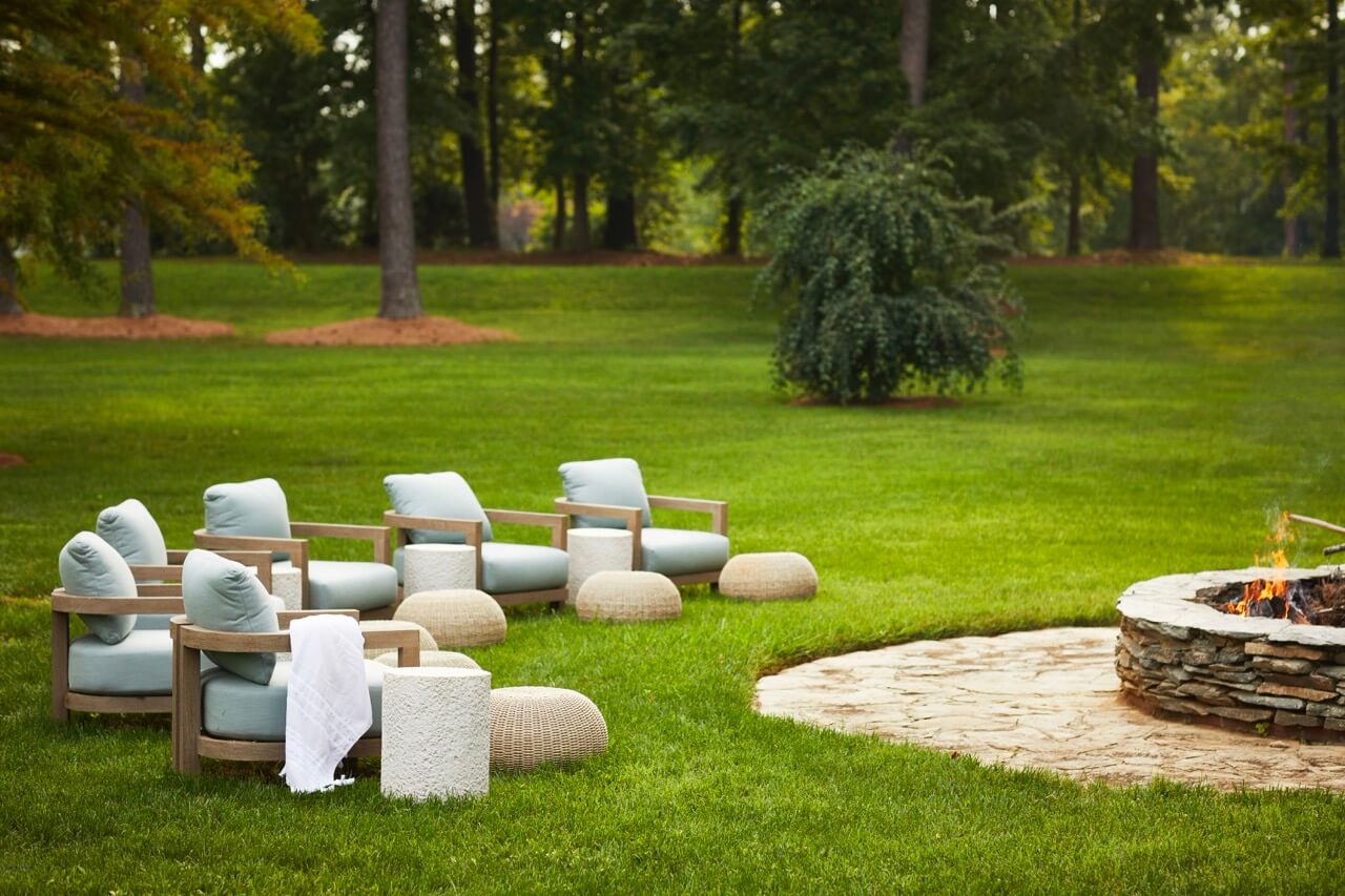 Four cushioned wooden chairs and woven poufs are arranged around a stone fire pit on a grassy lawn, creating the perfect spot to soak in lakeside beauty at this inviting South Carolina retreat, with trees framing the tranquil scene.