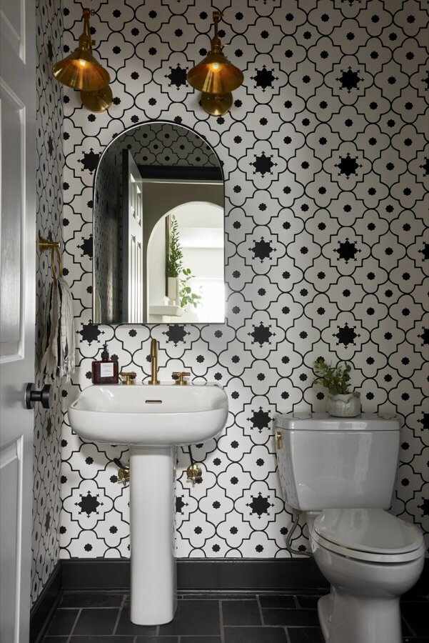 Bathroom with black-and-white patterned tile walls, a white pedestal sink, arched mirror, gold fixtures, toilet, and two brass pendant lights—an elegant example of interior design inspired by Silo Studio.