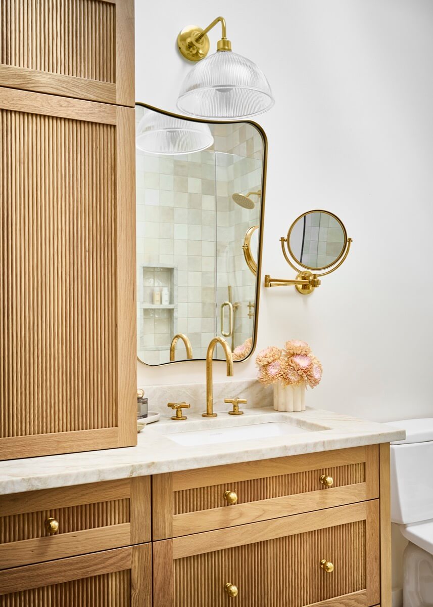 A bathroom vanity by Silo Studio features light wood cabinetry, a marble countertop, gold fixtures, a large mirror, and a wall-mounted lamp. A vase of pale flowers accents the serene interior design; a tiled shower is visible in the background.