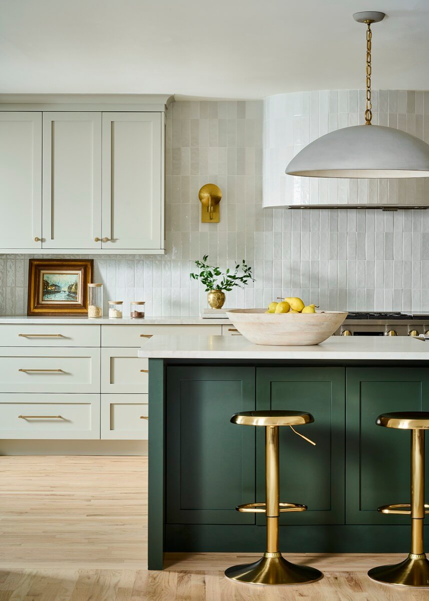 Modern kitchen by Silo Studio with white upper cabinets, green island, gold hardware, two gold stools, a bowl of lemons, and light wood flooring. 