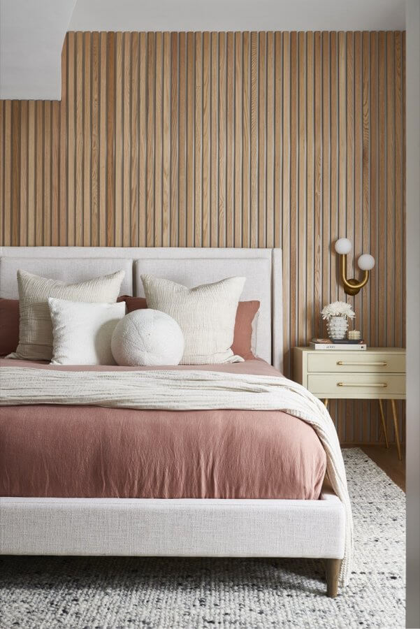 A modern bedroom by Silo Studio features a beige upholstered bed, rust-colored bedding, decorative pillows, a wood slat accent wall, and a cream nightstand with a gold lamp.