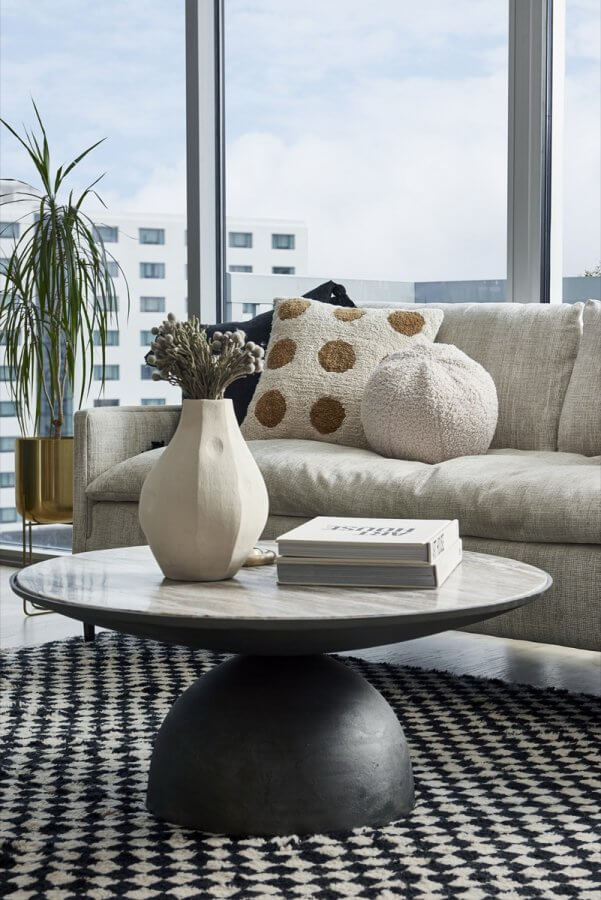 A modern Silo Studio living room featuring a beige sofa, textured cushions, a round coffee table with a vase and books, a patterned rug, and large windows framing city buildings.
