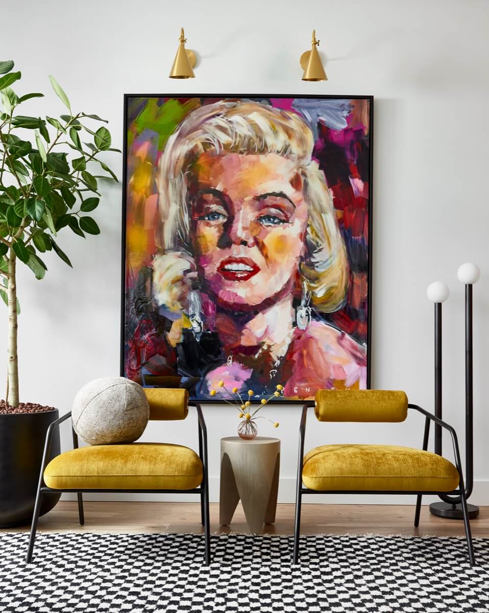 A modern living room by Silo Studio features two yellow chairs, a small side table, a potted plant, and a large colorful portrait of a woman on the wall—showcasing elegant interior design.
