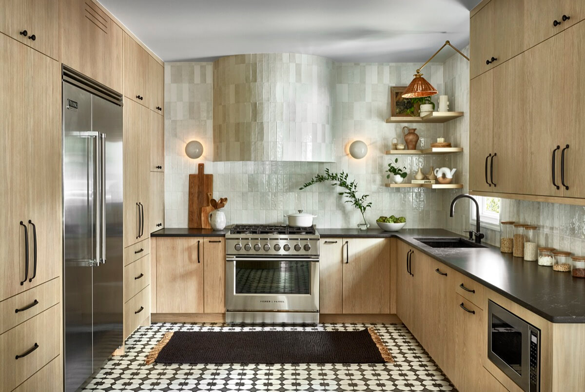 Modern kitchen with light wood cabinets, stainless steel appliances, black countertops, patterned tile floor, and white tiled backsplash. Floating shelves hold dishes and plants—an inviting space by Silo Studio.