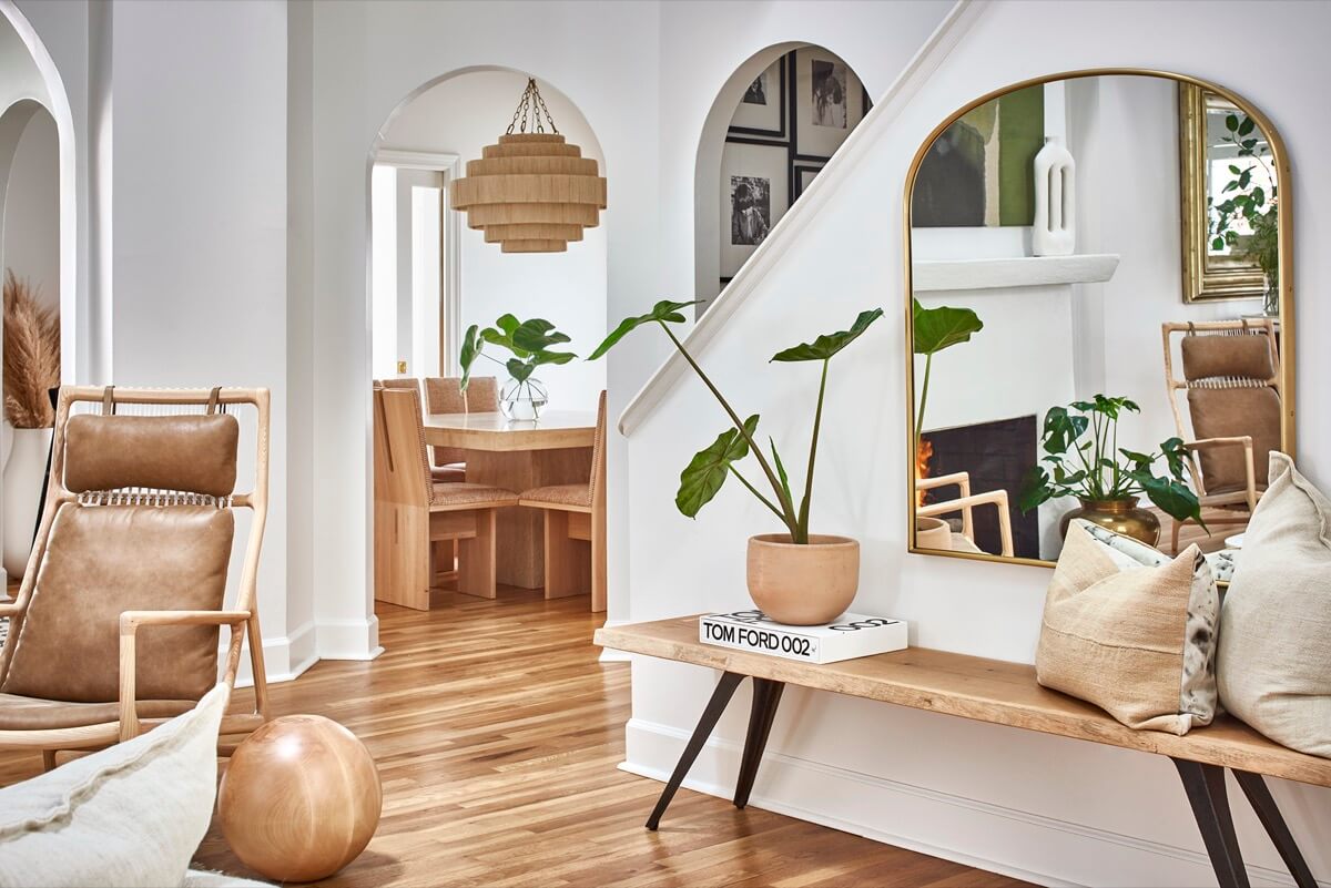 A modern living space designed by Silo Studio features wooden furniture, neutral tones, a potted plant on a bench, a large mirror, and a stylish dining area with pendant lighting.