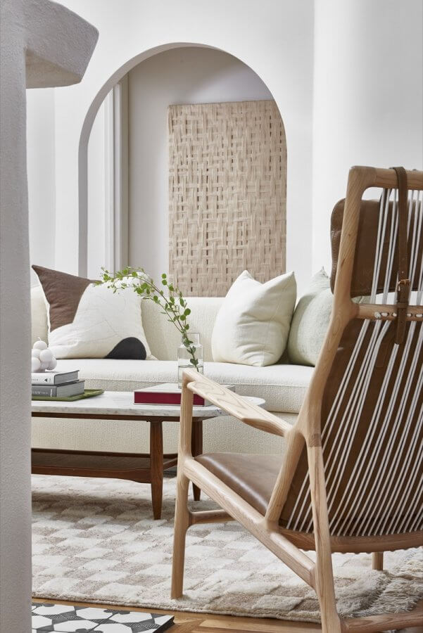 A modern living room by Silo Studio features a light sofa, neutral pillows, a wooden coffee table, a woven chair, and a decorative screen against a white wall with an arched doorway.