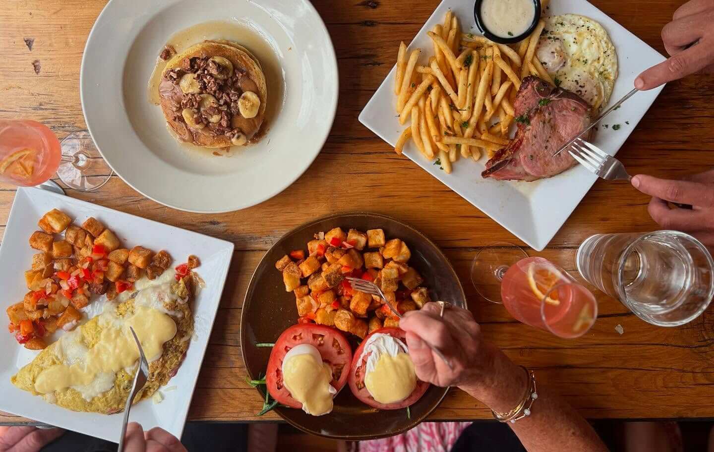 Overhead view of a brunch table with pancakes, eggs benedict, an omelette with potatoes, fries with ham and eggs, and two pink cocktails. Three people are seated, two holding forks.