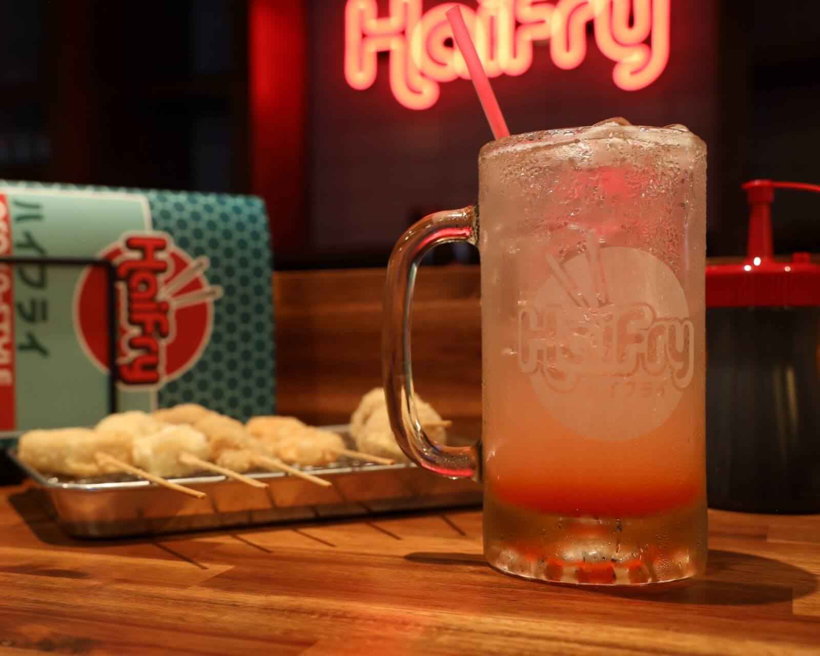 A frosty glass of a fizzy drink with a straw sits on a wooden table next to a tray of skewered fried food, with a branded box and neon sign in the background.