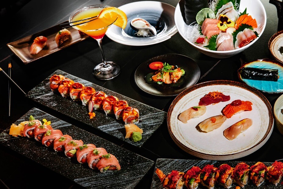 A table set with assorted sushi rolls, nigiri, sashimi, a cocktail, and various Japanese appetizers served on black and white plates.