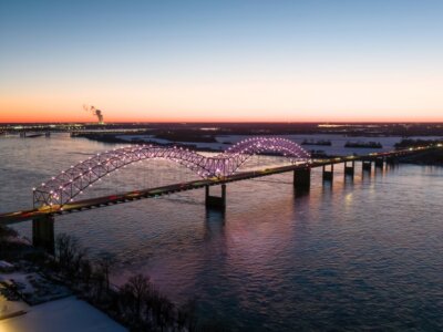 Aerial view of a large, illuminated arch bridge spanning a wide river at dusk, with traffic crossing and city lights in the distance—capturing the vibrant atmosphere that surrounds Memphis events in August.