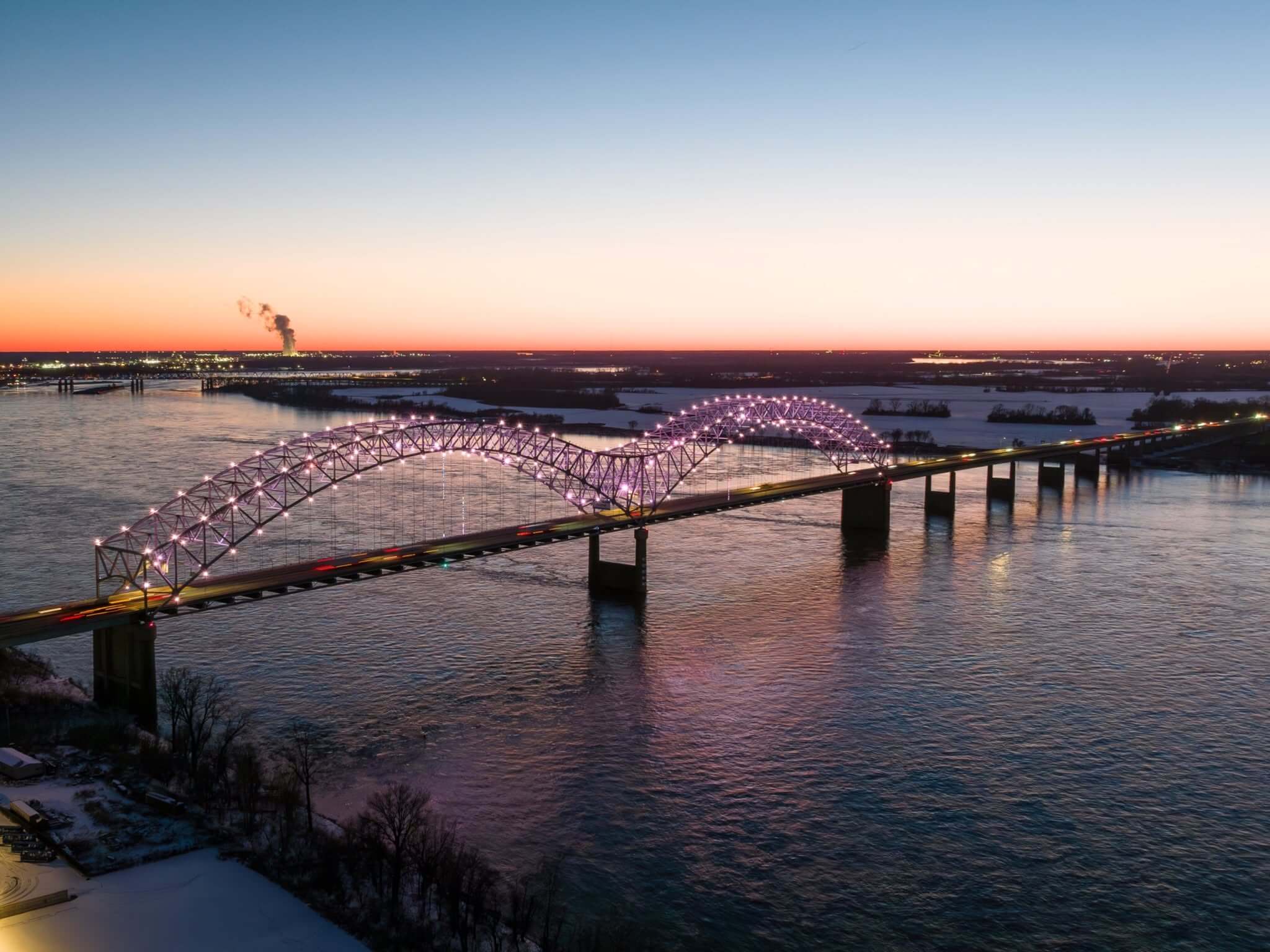 Aerial view of a large, illuminated arch bridge spanning a wide river at dusk, with traffic crossing and city lights in the distance.