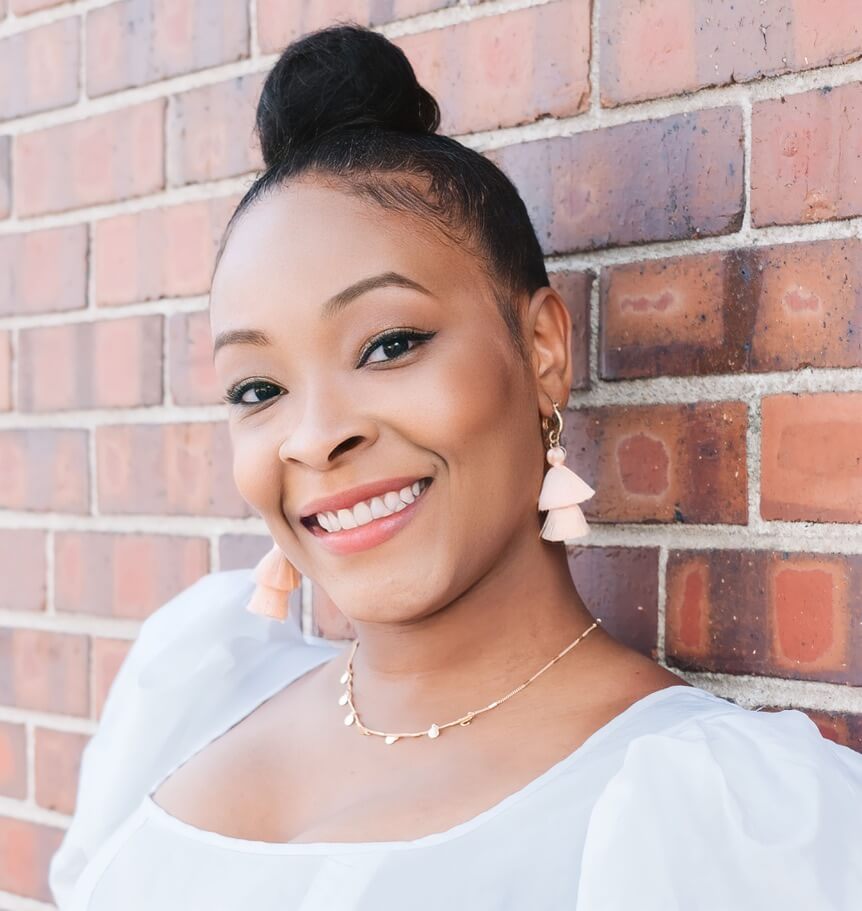 A woman with a top bun hairstyle, wearing tassel earrings and a white blouse, smiles while leaning against a brick wall.