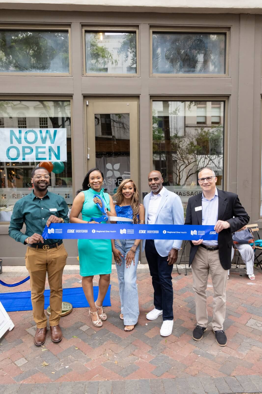 Five adults, including Antwanette Crutcher, stand outside a storefront with a "Now Open" sign, holding a blue ribbon for the FACES ribbon-cutting ceremony.