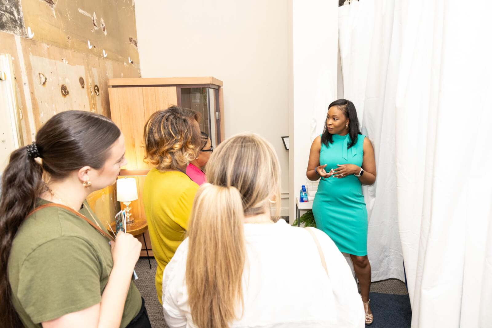Antwanette Crutcher, in a teal dress, speaks to four people in a room with white curtains and unfinished walls.