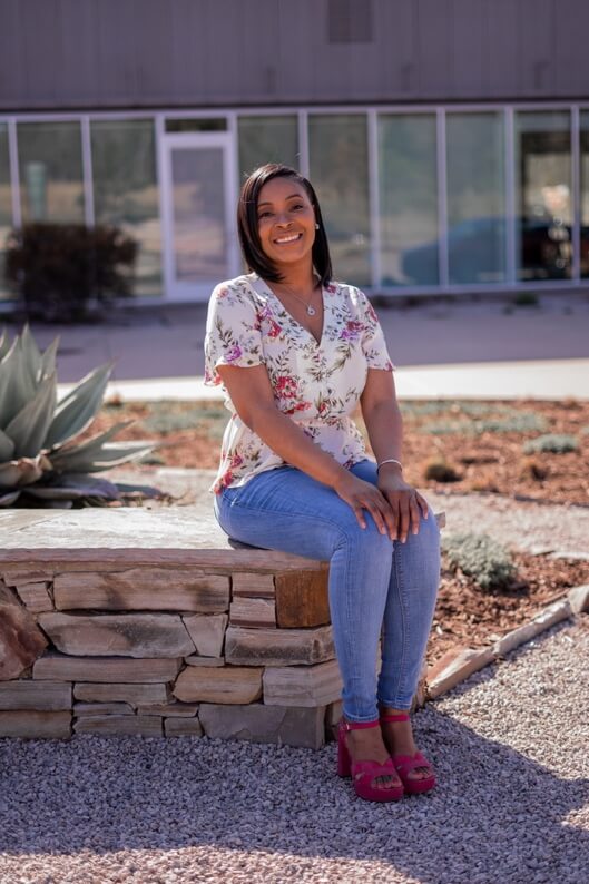A woman wearing a floral blouse, jeans, and pink shoes sits on a stone bench outdoors, smiling at the camera—radiating the serenity of an urban wellness retreat.