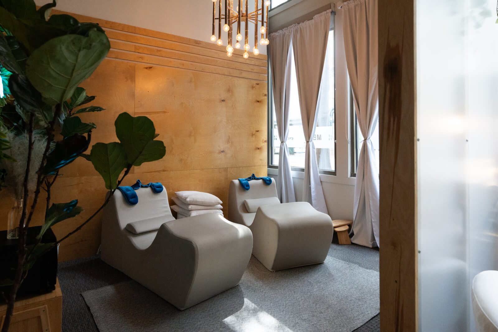 Two modern lounge chairs with blue towels and pillows face a window with white curtains in a softly lit, contemporary relaxation room.