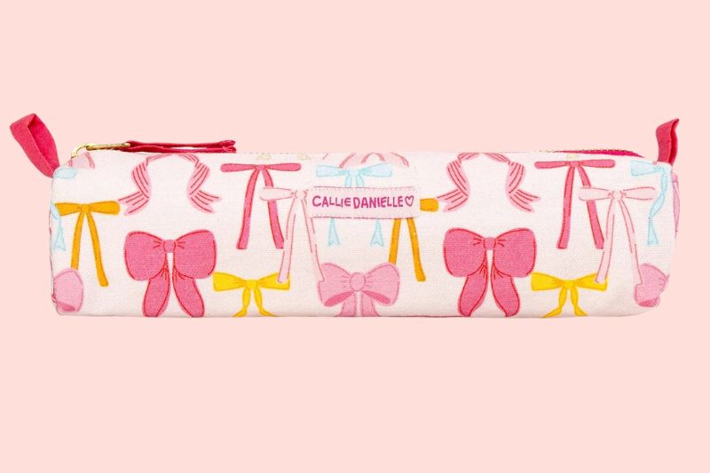 A rectangular pencil case with a pink zipper, decorated with pink, yellow, and blue bow patterns on a light pink background—a cute Memphis FINDS for August 2025.