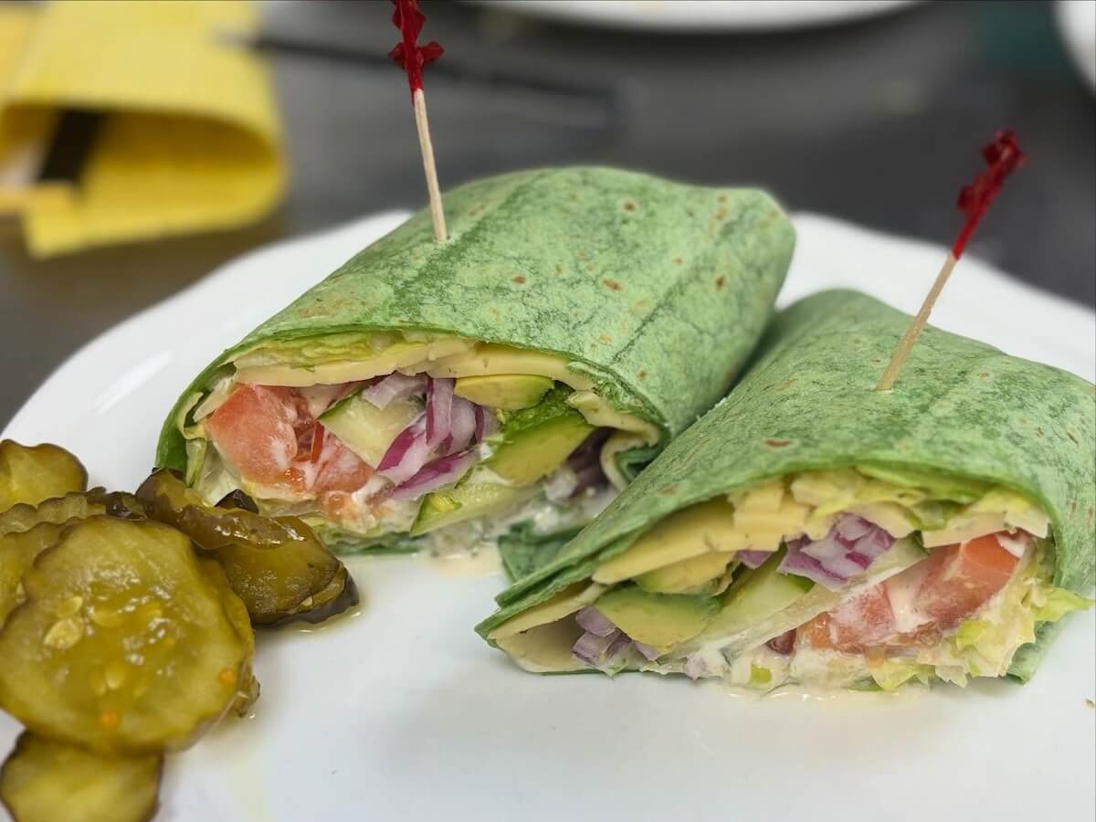 A spinach wrap filled with lettuce, tomato, avocado, red onion, and cheese is cut in half and served on a white plate with sliced pickles.