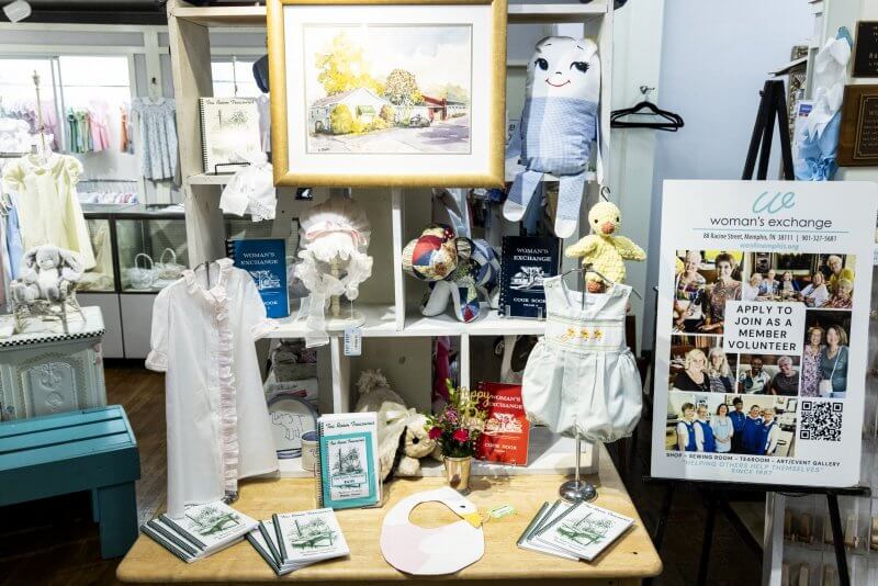 A display table with vintage baby clothes, stuffed toys, books, framed art, and a sign inviting the community to join or volunteer at the Woman’s Exchange of Memphis.