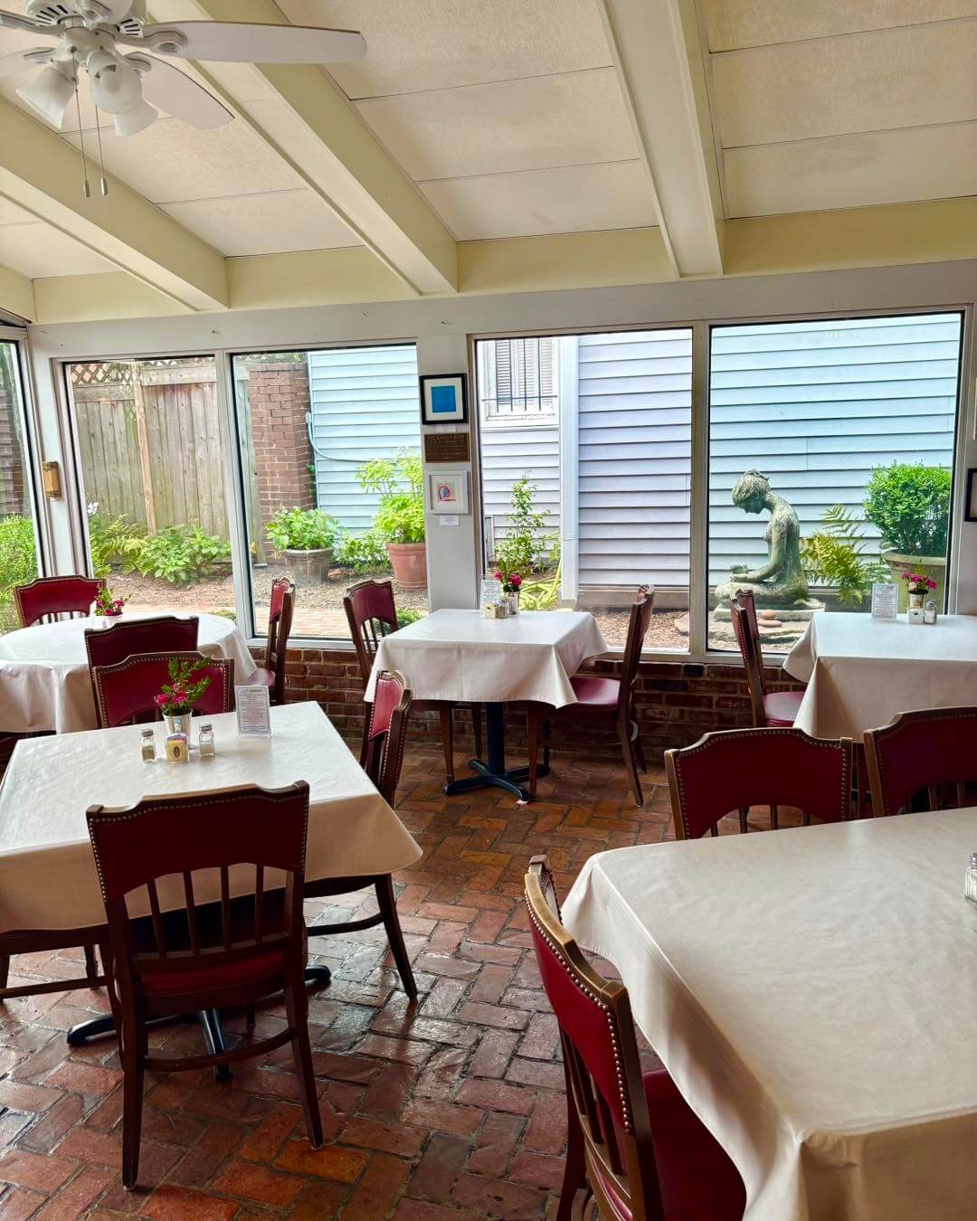 A small dining area with tables and chairs, brick floor, and large windows overlooking a garden with a statue—perfect for fostering community at the Woman’s Exchange of Memphis.