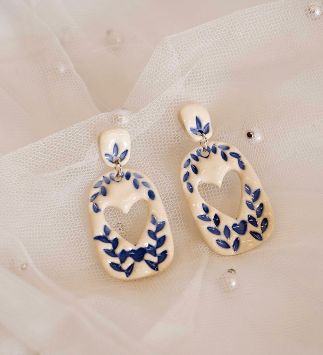 18 Nashville FINDS for Late Summer: Jewelry, Decor & More - 17 A pair of white ceramic earrings with blue leaf designs and heart-shaped cutouts, perfect for your Late Summer FINDS in Nashville, placed on sheer fabric with small pearls.