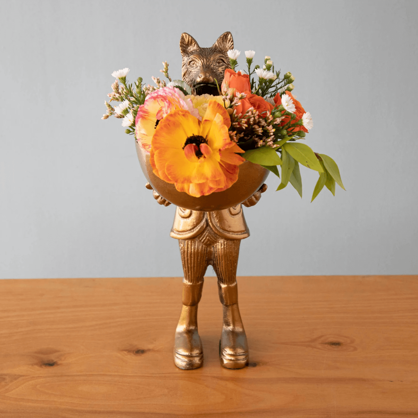 18 Nashville FINDS for Late Summer: Jewelry, Decor & More - 5 A gold figurine with an animal head holds a bowl of assorted orange, yellow, and white flowers—perfect for Nashville-inspired Late Summer FINDS—standing on a wooden surface against a plain gray background.