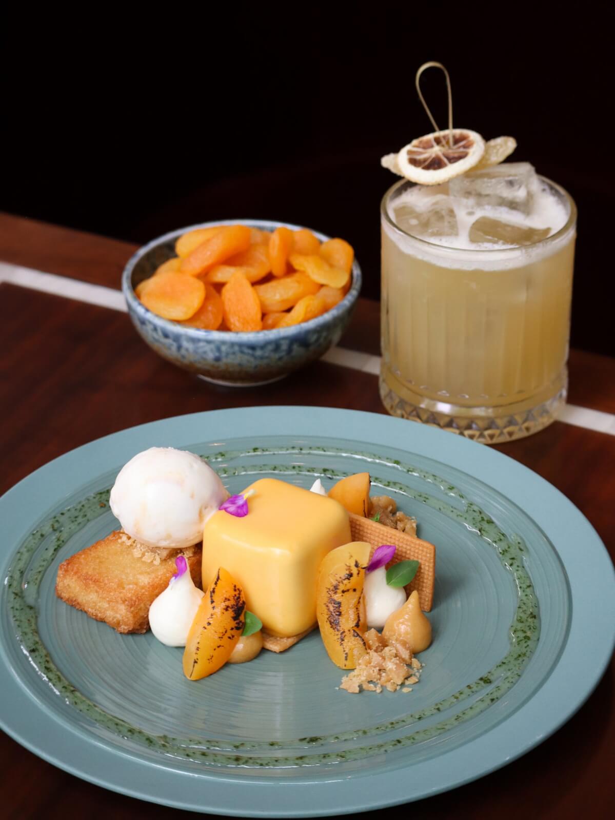 A plated dessert with a yellow mousse cube, grilled fruit, ice cream, and garnishes, accompanied by a cocktail and a bowl of dried apricots on a wooden table—perfect for savoring sweet Nashville deals.