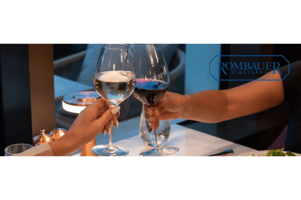 Two people clinking glasses of white and red wine at a dining table with the Rombauer Vineyards logo in the background.