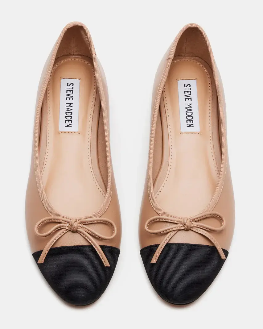 A pair of beige ballet flats with black capped toes, bow accents, and "Steve Madden" labels on the insoles, embodying French girl style, photographed from above on a white background.
