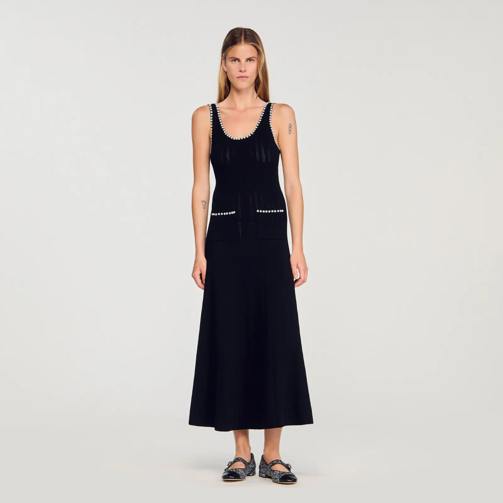 A woman stands against a plain background in a sleeveless black knit top and matching midi skirt, paired with black loafers—an embodiment of effortless style.