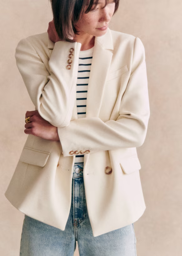 A person channels French Girl Style, wearing a cream-colored blazer over a black and white striped shirt and light blue jeans, standing against a neutral background.