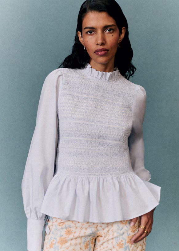 Woman with long dark hair wearing a white long-sleeve blouse with ruffled details and light-patterned pants, showcasing effortless style against a plain blue background.