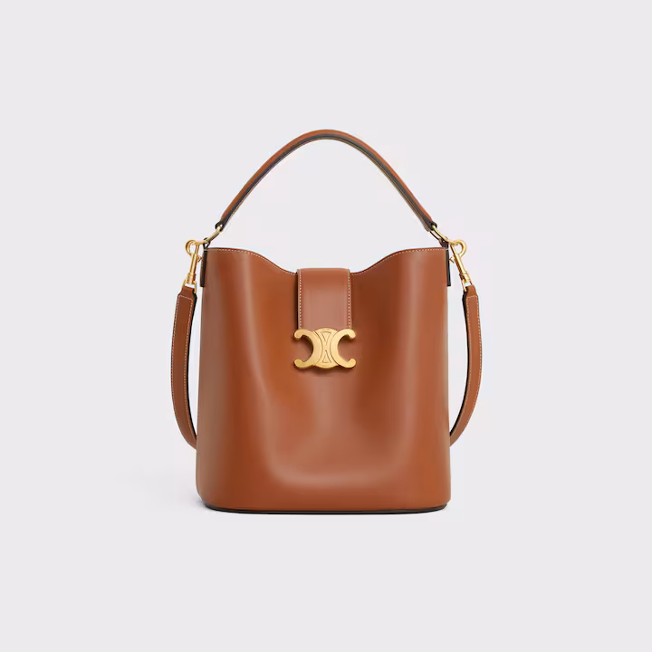 Brown leather bucket bag with a top handle, detachable shoulder strap, and gold-tone hardware featuring a decorative logo on the front—perfect for capturing that effortless French girl style.