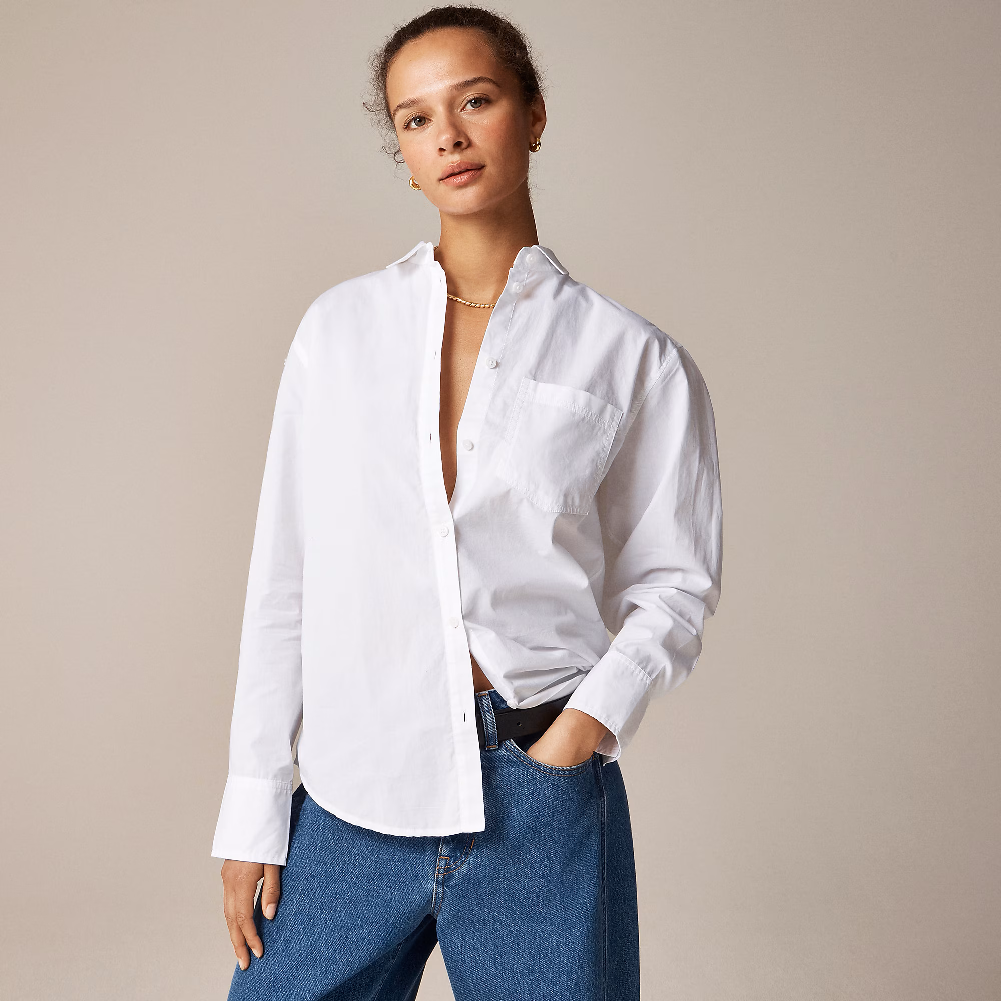 Person wearing a white button-up shirt with the top buttons undone, blue jeans, and a necklace, standing against a plain beige background, channeling effortless style inspired by French fashion.