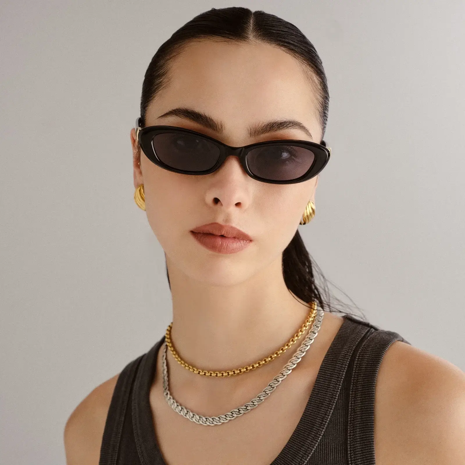 Woman with dark hair in a sleek ponytail wearing black oval sunglasses, gold hoop earrings, layered chain necklaces, and a sleeveless dark top—an effortless style that channels classic French girl style.