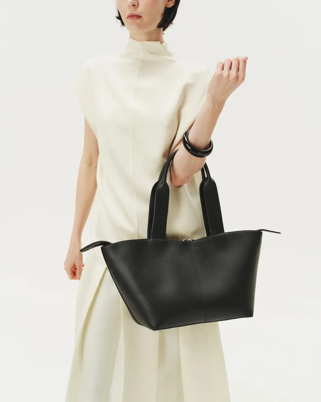 A person in a white dress showcases effortless style, holding a large black leather handbag with wide handles while standing against a plain white background.