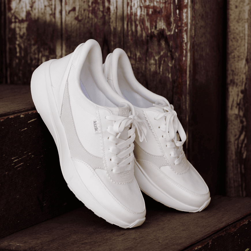 A pair of white athletic sneakers with laces resting on dark wooden stairs, positioned against a weathered wooden wall—capturing effortless style with a hint of French girl style charm.