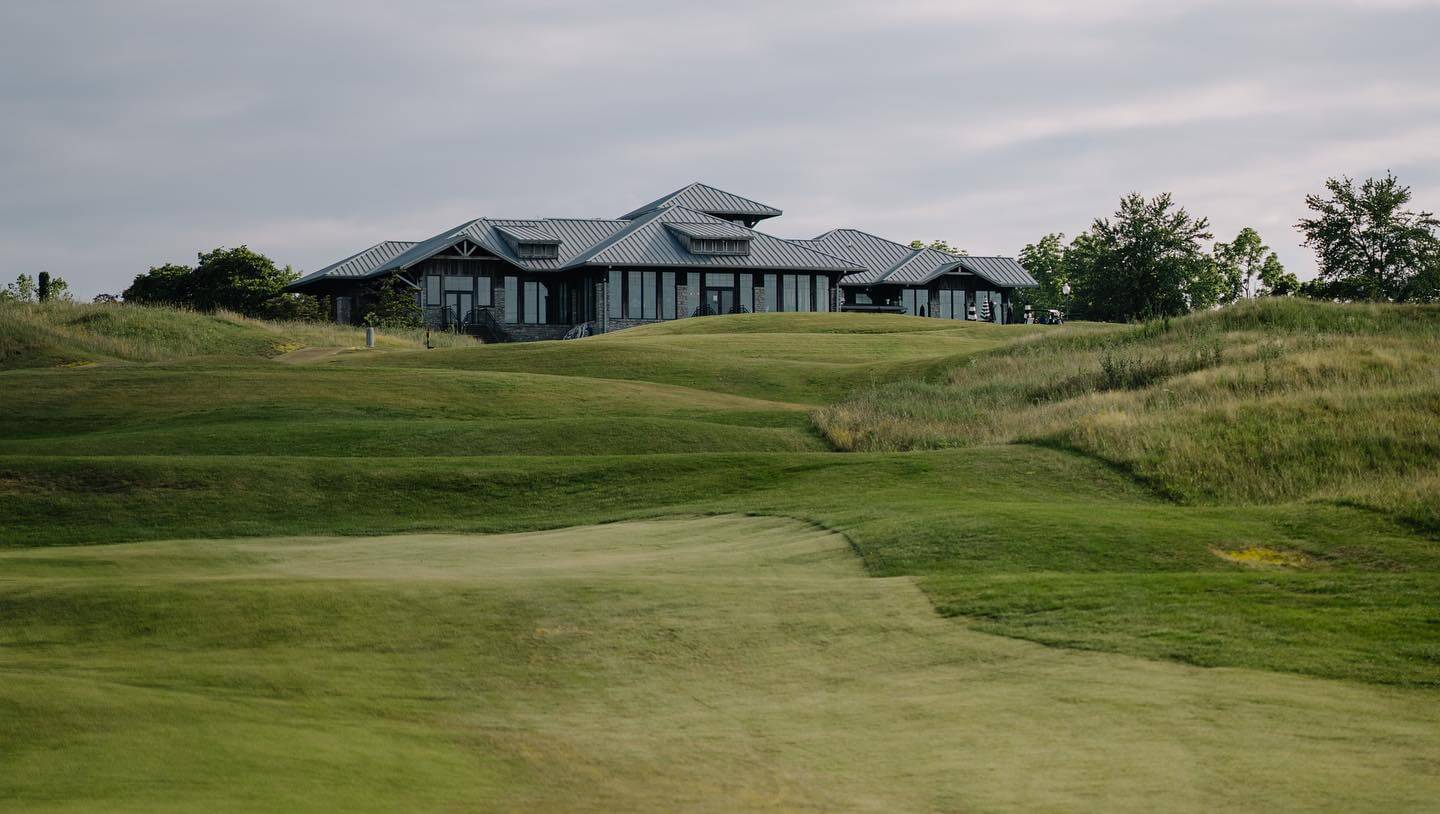 A modern building with large windows sits atop rolling green hills on a golf course under a cloudy sky, showcasing the scenic beauty found at premier Nashville golf courses.