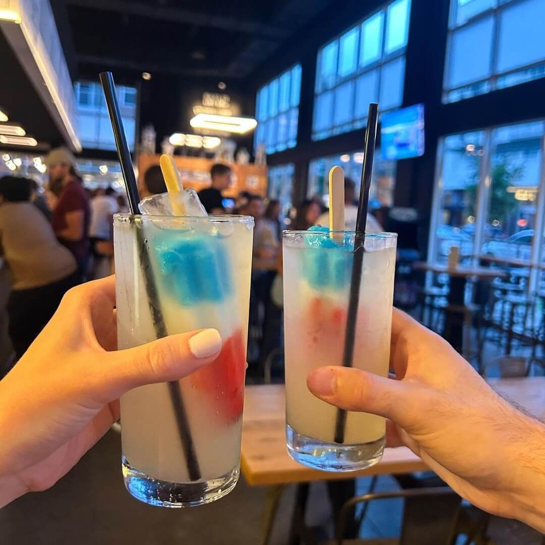 Two hands hold up tall glasses of lemonade with black straws and red, white, and blue popsicles inside, in a modern bar or restaurant with large windows—perfect for relaxing after a fun night at bars with games.