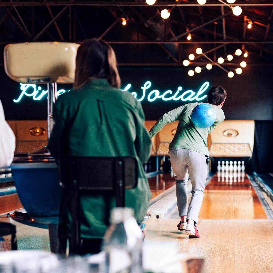 A person bowls a blue ball at a bowling alley—one of the lively bars with games—while another sits nearby; a neon sign reads "Pinewood Social" in the background.