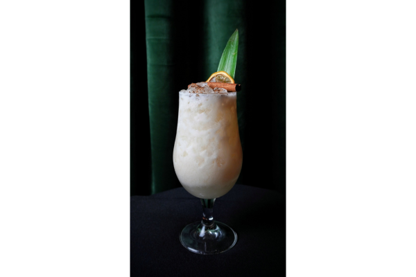 A creamy Tiki Cocktail served in a stemmed glass, garnished with a dried orange slice, pineapple leaf, and cinnamon stick, set against a dark green background—perfect for the Masters Series exploring advanced techniques.